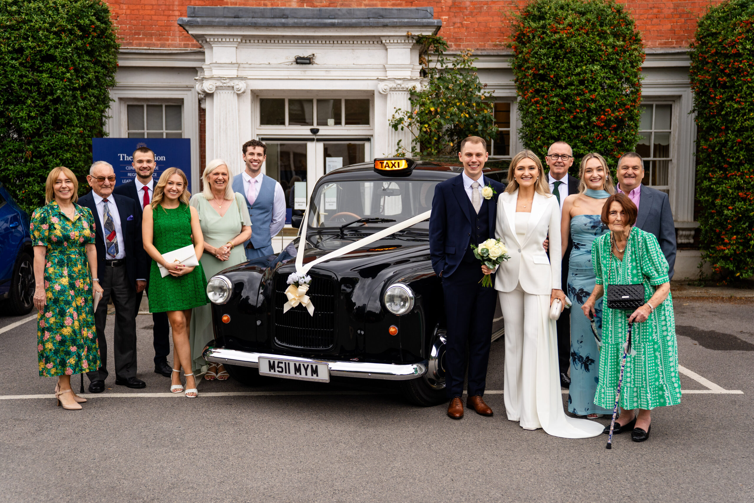 Bride and groom in the front of Leatherhead registry office with taxi with family